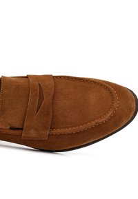 Mocasin de gamuza marrón con punta redondeada, que presenta una costura y una banda decorativa en la parte superior. Textura suave y suela flexible.