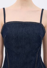 Denim-Oberteil mit quadratischem Ausschnitt, dünnen Trägern und kontrastierender gelber Naht. Der Stoff ist dunkelblau und hat eine glatte Textur.