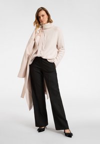 Hellrosa gerippter Rollkragenpullover, schwarze weite Hosen und ein langer beigefarbener Mantel, ergänzt durch schwarze Stilettos mit Schleifenakzenten.