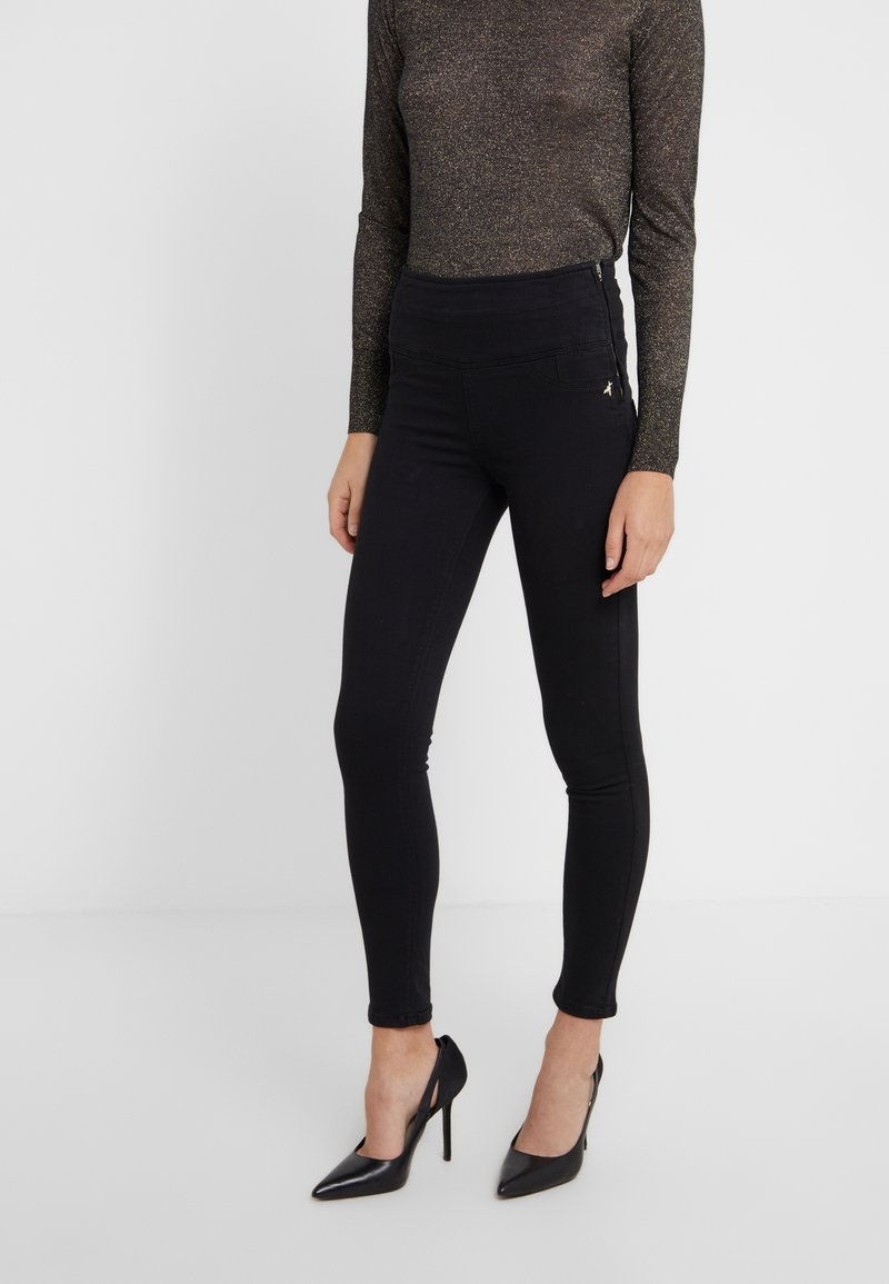 Femme portant un pantalon skinny noir taille haute, un haut marron transparent à manches longues et des talons hauts noirs pointus, debout sur un fond blanc uni.