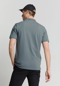 Grijze polo met korte mouwen en een klassieke kraag, met een gladde textuur en zijsplitten. Een zwarte pet met een gebogenrand maakt de outfit compleet.