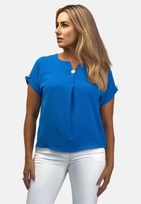 Niebieska bluzka z krótkim rękawem, z okrągłym dekoltem i akcentującym guzikiem, wykonana z teksturowanej tkaniny, zestawiona z białymi spodniami.