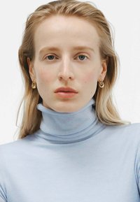 Suéter de cuello alto azul claro con un diseño ajustado. Pendientes en forma de lágrima dorados acentúan el rostro de la modelo y llaman la atención sobre el escote.