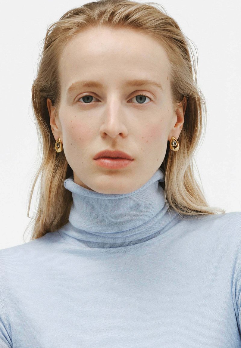 Suéter de cuello alto azul claro con un diseño ajustado. Pendientes en forma de lágrima dorados acentúan el rostro de la modelo y llaman la atención sobre el escote.