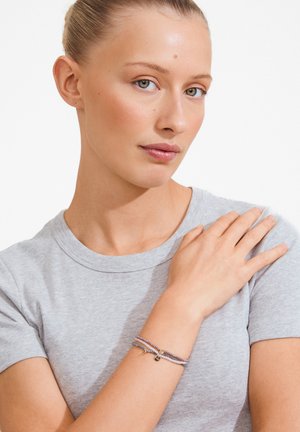 Mladá žena v šedom tričku s vlasmi stiahnutými dozadu, nosí viacvrstvové korálkové náramky na zápästí a odpočíva rukou na opačnom pleci.