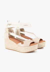 Sandalias de cuña de espadrille beige con una suela trenzada texturizada, punta cerrada y correas en la parte del tobillo entrelazadas. Acentuadas con un brillo metálico.