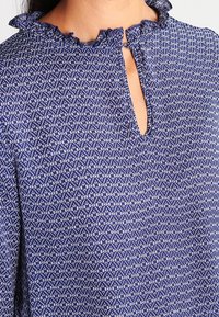 Blaue Bluse mit einem Rüschenausschnitt und einem kleinen Schlüsselloch. Der Stoff hat ein strukturiertes Design in einem geometrischen Muster.