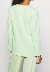 Jasnozielony, fakturowany top z długim rękawem o prostym kroju i bocznymi rozcięciami, noszony z jasnozielonymi spodniami w kropki.