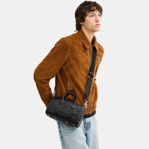 Mladić u smeđoj jakni od brušene kože i svijetloplavim trapericama, nosi crnu uzorkovanu kožnu torbu s remenom preko ramena.