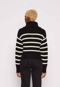 Person mit kurzen lockigen blonden Haaren, die einen schwarz-weiß gestreiften Pullover und schwarze Hosen trägt, mit dem Rücken zu einem einfarbigen hellen Hintergrund.