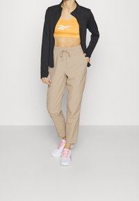 Kvinna i svart jacka, orange sportbH med vit logotyp, beige byxor med snörning och vita sneakers med rosa snören står på grått golv.