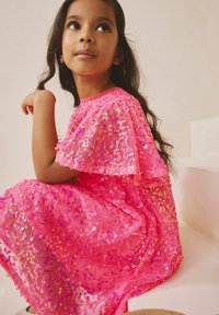 Rózsaszín flitteres ruha lebegő ujjakkal, áttetsző réteggel és különböző árnyalatú rózsaszín flitterekkel, amelyek csillogó textúrát hoznak létre.