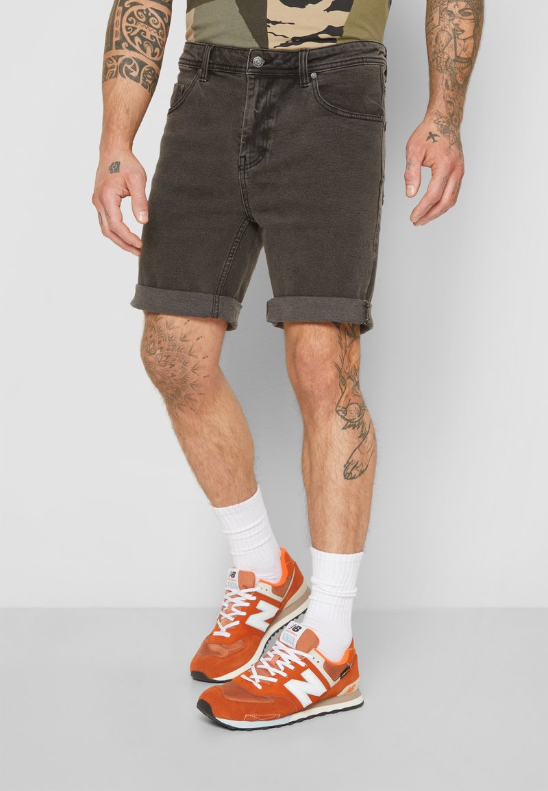 Tumši pelēki denima šorti ar sarullētiem maliņām, apvienoti ar oranžiem un baltajiem New Balance sporta apaviem. Teksturēta virsma, redzamās šuvju detaļas un kabatas.