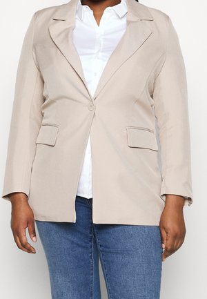 Femme portant un blazer beige à un bouton sur une chemise blanche et un jean bleu, vue du cou aux hanches sur un fond neutre.