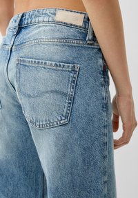 Primer plano de unos jeans de mezclilla azul claro con un bolsillo trasero y una etiqueta de marca beige, llevados por una persona con el abdomen desnudo y el brazo visible.
