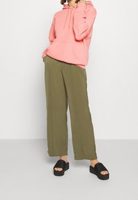 Korálově růžová mikina s kapucí a přední kapsou, doplněná olivově zelenými širokými kalhotami a černými platformovými sandály s proužkovanými detaily.