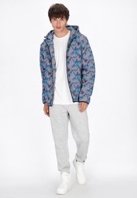 Giacca a vento da uomo con un motivo geometrico colorato in blu, rosso e bianco, abbinata a pantaloni tuta grigi e sneaker bianchi.