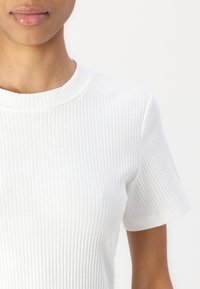 Hvit ribbet t-skjorte med rund hals og korte ermer. Stoffet har en glatt tekstur og en tettsittende form som definerer silhuetten.