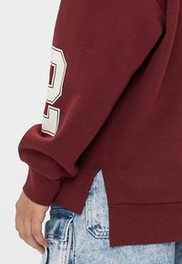 Sweatshirt bordô com decote redondo e punhos canelados. Apresenta o número 92 impresso em branco na manga esquerda. Abertura lateral na bainha.