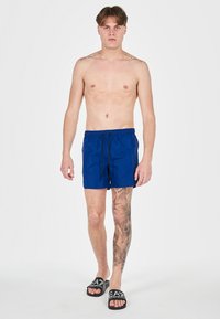 Costumi da bagno blu con vita elastica e lacci, caratterizzati da una trama liscia. Il modello indossa ciabatte nere. Il tono della pelle è chiaro.