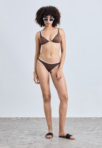 Brown bikini with white trim, featuring a triangle top with a front knot detail. Model wears black textured slides and white cat-eye sunglasses.