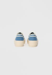 Plave platnene tenisice s bež potplatom i naglašenim prugama. Imaju nisku siluetu, zaobljeni vrh i teksturiranu tkaninu.