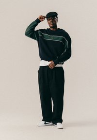 Tumedat ja rohelised triibulised dressipluusid vabamises lõikes, koos mustade pükste ja valgete Nike tennistega. Mudel kannab lame peakattet ja prille.