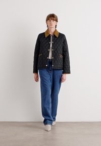 Jackett negru matlasat cu guler maro, accesorii aurii și buzunare laterale, asortat cu blugi largi albaștri și pantofi bej.