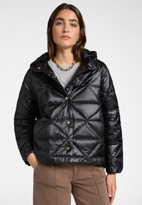 Chaqueta de plumas negra con capucha, con un diseño de acolchado en diamante, botones de presión y un acabado brillante. Llevada sobre un suéter gris.