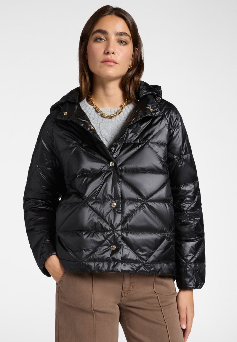 Chaqueta de plumas negra con capucha, con un diseño de acolchado en diamante, botones de presión y un acabado brillante. Llevada sobre un suéter gris.