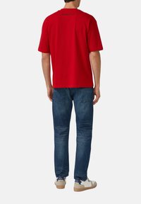 Rotes Kurzarm-T-Shirt mit Rundhalsausschnitt, mit Logodruck auf dem Rücken. Kombiniert mit blauen Jeans und weißen Sneakers.