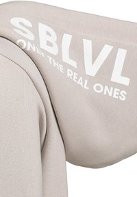Beige Kapuzenpullover aus strukturiertem Material. Der Ärmel zeigt weißen Drucktext "SBLVL ONLY THE REAL ONES." Schlichtes Design und lockere Passform.