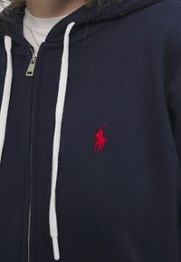 Marinblå ziptröja med vita dragsnören, prydd med en liten röd broderad polo-spelare logo på vänster bröst. Tillverkad av en bomullsblandning.