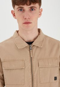 Beige glidelås overskjorte laget av solid stoff, med to frontlommer og et lite logo-påse på høyre lomme.