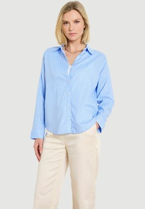 Žena v svetlomodrej pruhovanej košeli na gombíky a krémových nohaviciach stojaca s jednou rukou v vrecku na jednoduchom pozadí.