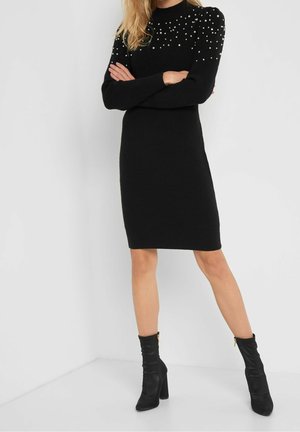 Donna che indossa un abito maglione nero al ginocchio con decorazioni di perle sulle spalle, abbinato a stivaletti neri con tacco.