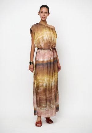 Femme portant une robe maxi asymétrique tie-dye dans des teintes marron, jaune et rose avec une fine ceinture, des sandales marron tressées et un bracelet manchette métallique.