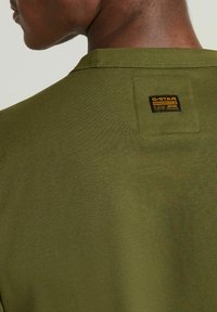 Ingrandimento di una persona che indossa una camicia verde oliva con un piccolo etichetta nera e gialla G-Star Originals Raw Denim sulla parte superiore della schiena.
