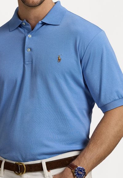 Svetlo modra polo majica iz bombaža. Ima klasičen ovratnik, tri gumbe in izvezen logo. Oblečena z rjavim pasom in uro.