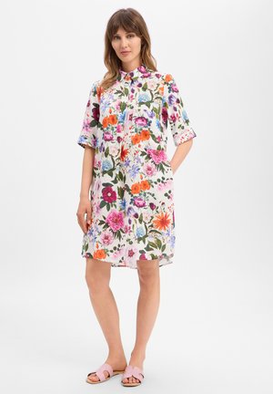 Vrouw draagt een knielange, korte mouwen, bloemendessin jurk met kraag en roze slidesandalen, staand met één hand in de zak.