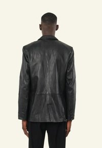 Zwarte leren blazer met een licht getextureerde afwerking, voorzien van een klassieke kraag, rugventilatie en knoopsluiting bij de manchetten, gezien van de achterkant.