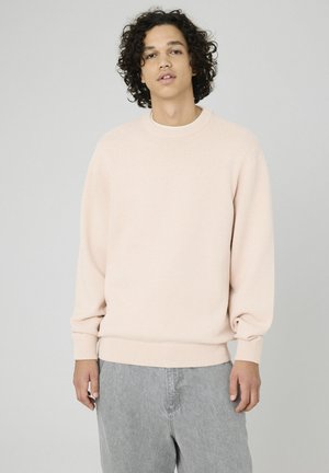 Pull en maille rose clair avec un col rond, des poignets et un ourlet côtelés. Texture douce, coupe décontractée. Porté sur un jean gris.