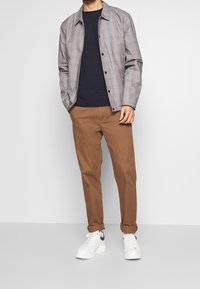 Muž ve šedé kostkované košili s knoflíky přes černé tričko, hnědých vyhrnutých kalhotách a bílých teniskách s černým logem Nike.