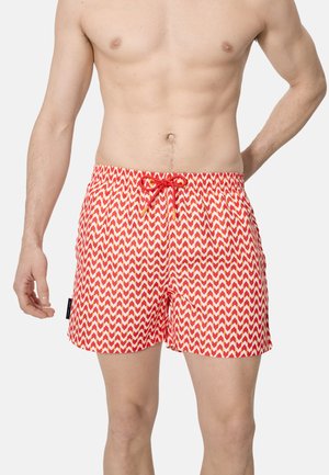 Hombre sin camisa que lleva pantalones de baño rojos y blancos con patrón de zigzag y cordón rojo, de pie con una mano apoyada en la cadera.