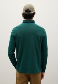 Langarm-Polohemd aus dunkelgrünem Baumwollstoff mit einem navyfarbenen Kragen. Die Person trägt khakifarbene Hosen und eine Hundezahnmuster-Mütze.