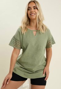 Olivgrön topp med texturerad tyg, nyckelhålshalsslinning och korta ärmar av spets. Tillsammans med svarta shorts poserar modellen mot en ljus bakgrund.