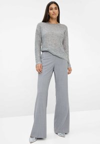Hellgrauer Pullover mit strukturiertem Strick und Schimmer, kombiniert mit grauen Hochbundstreifen-Hosen. Silberne spitze Schuhe runden das Outfit ab.