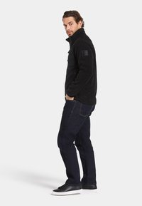 Svart fleecejacka med hög krage, dragkedjefront och bröstficka. I kombination med mörka jeans och svarta sneakers. Strukturerad yta.