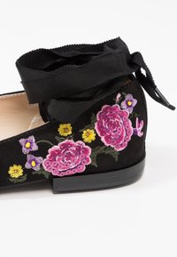 Chaussure plate noire avec rubans à nouer autour de la cheville, décorée de fleurs brodées roses, jaunes et violettes ainsi que de sequins sur fond blanc.