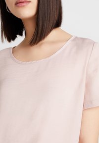 Ljust rosa kortärmad topp med rundad halsringning. Tyget verkar slätt med en lätt lyster. Enkel design, inga synliga mönster eller detaljer.